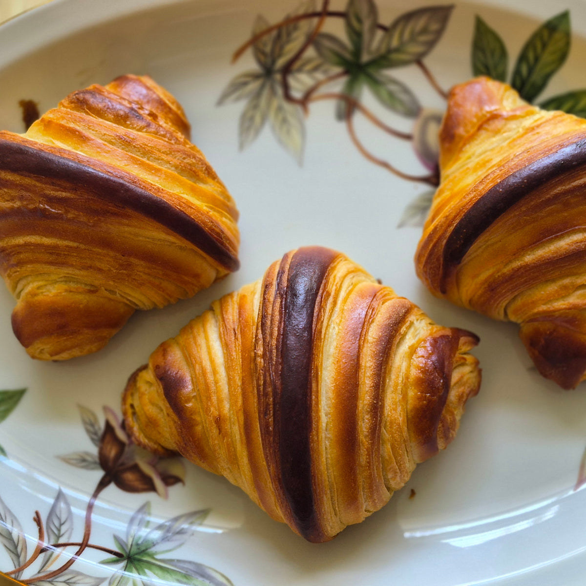 Three golden-brown croissants arranged on a white ceramic plate with a green and brown leaf pattern, set on a light wooden surface.