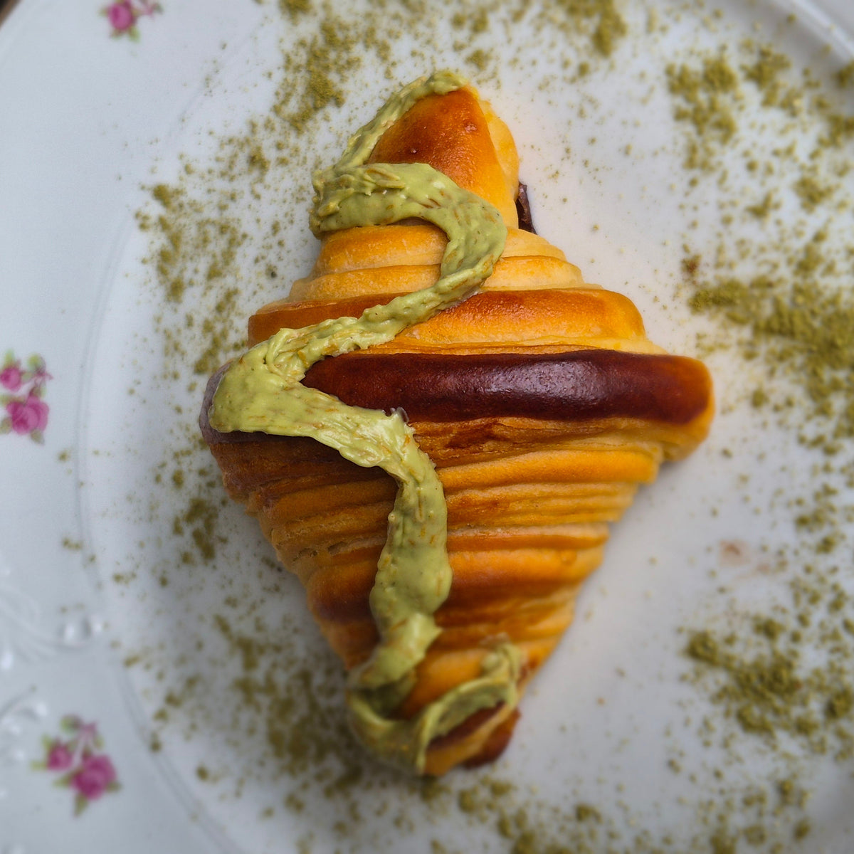 A close-up shot of a sweet pastry, likely a banana and chocolate croissant, drizzled with green matcha paste and dusted with matcha powder, served on a white floral ceramic plate.