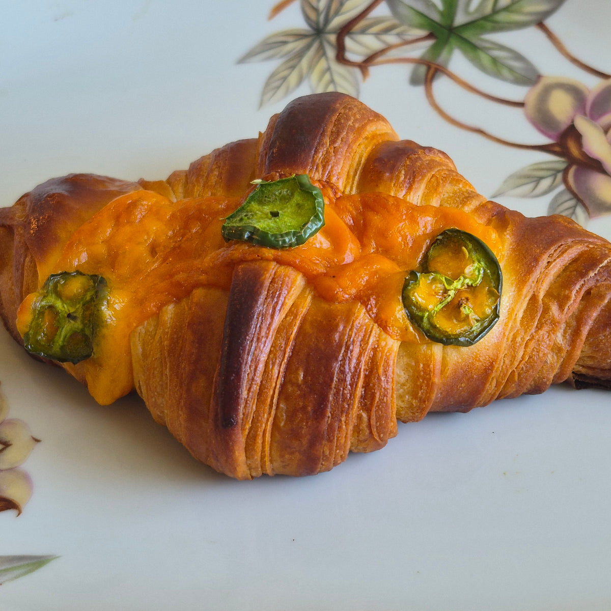 A baked croissant-style pastry with melted orange cheese and sliced jalapeño peppers, placed on a white floral surface.