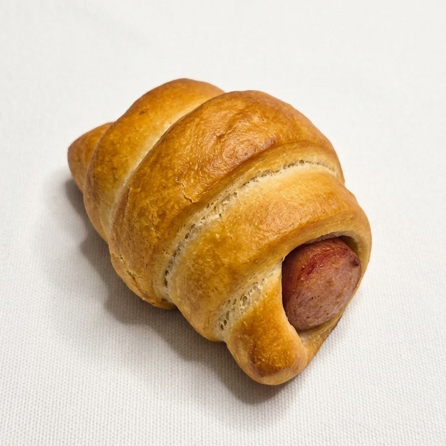A golden-brown baked pastry shaped like a croissant, filled with a small sausage slice, photographed on a plain light background.
