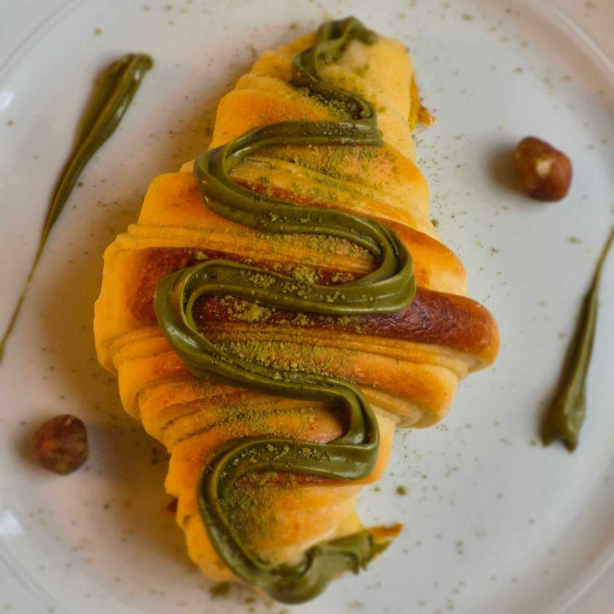 A close-up shot of a pastry croissant filled with green matcha cream and topped with matcha powder, served on a white plate with hazelnuts and drizzles of matcha cream.