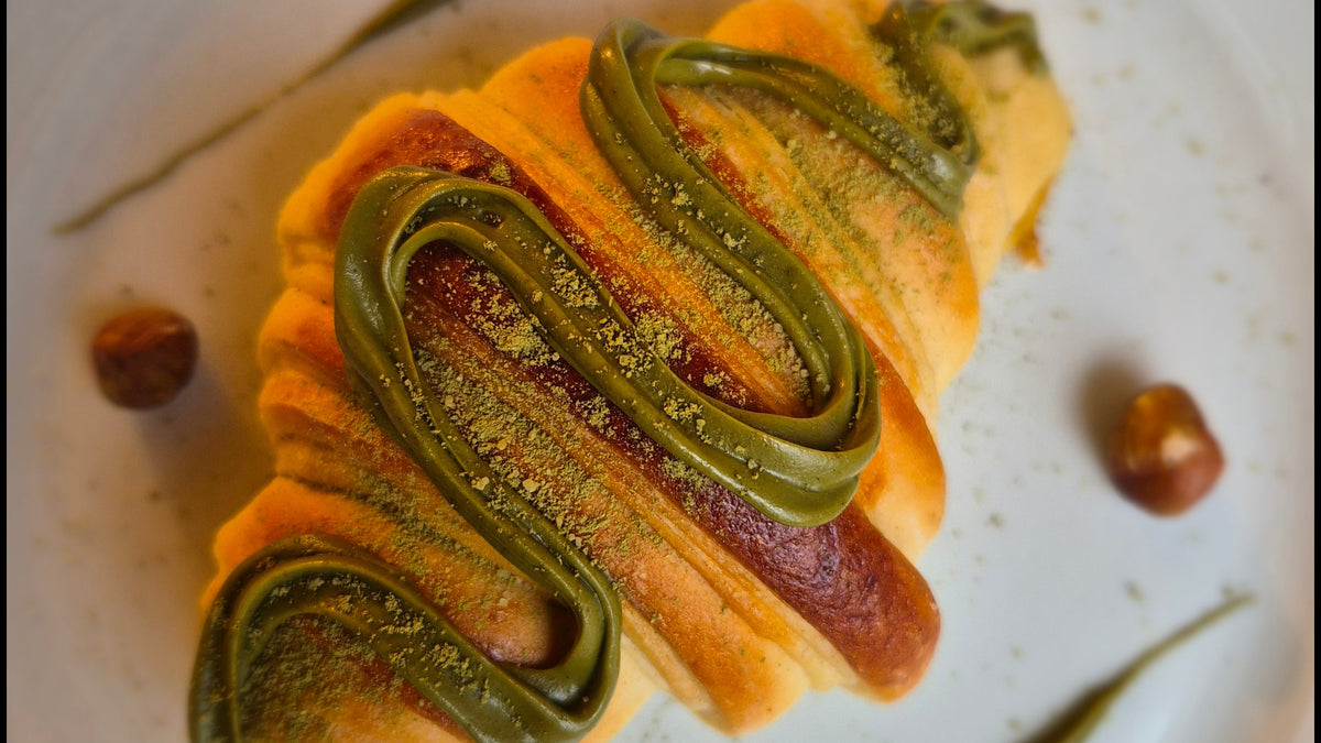 A close-up shot of a pastry croissant filled with red jam and drizzled with green matcha cream, garnished with a sprinkle of matcha powder, served on a white floral ceramic plate with hazelnuts.